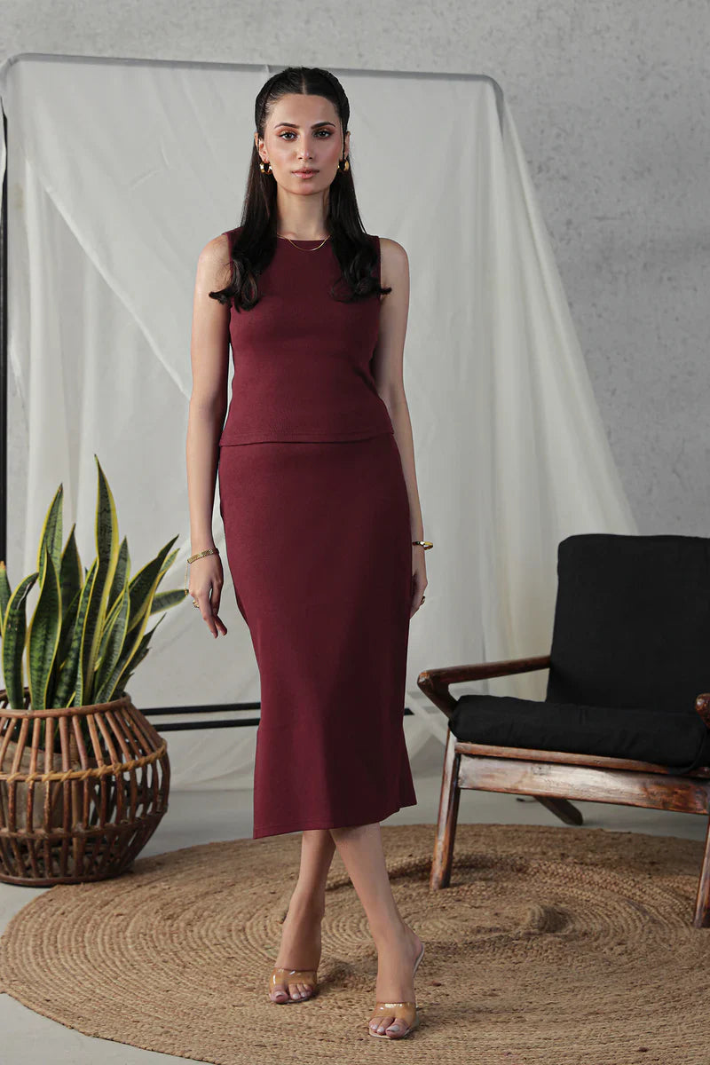A woman in a maroon sleeveless dress with a matching skirt stands in a minimalistic setting, featuring a plant in a woven basket, a wooden chair, and a white backdrop.