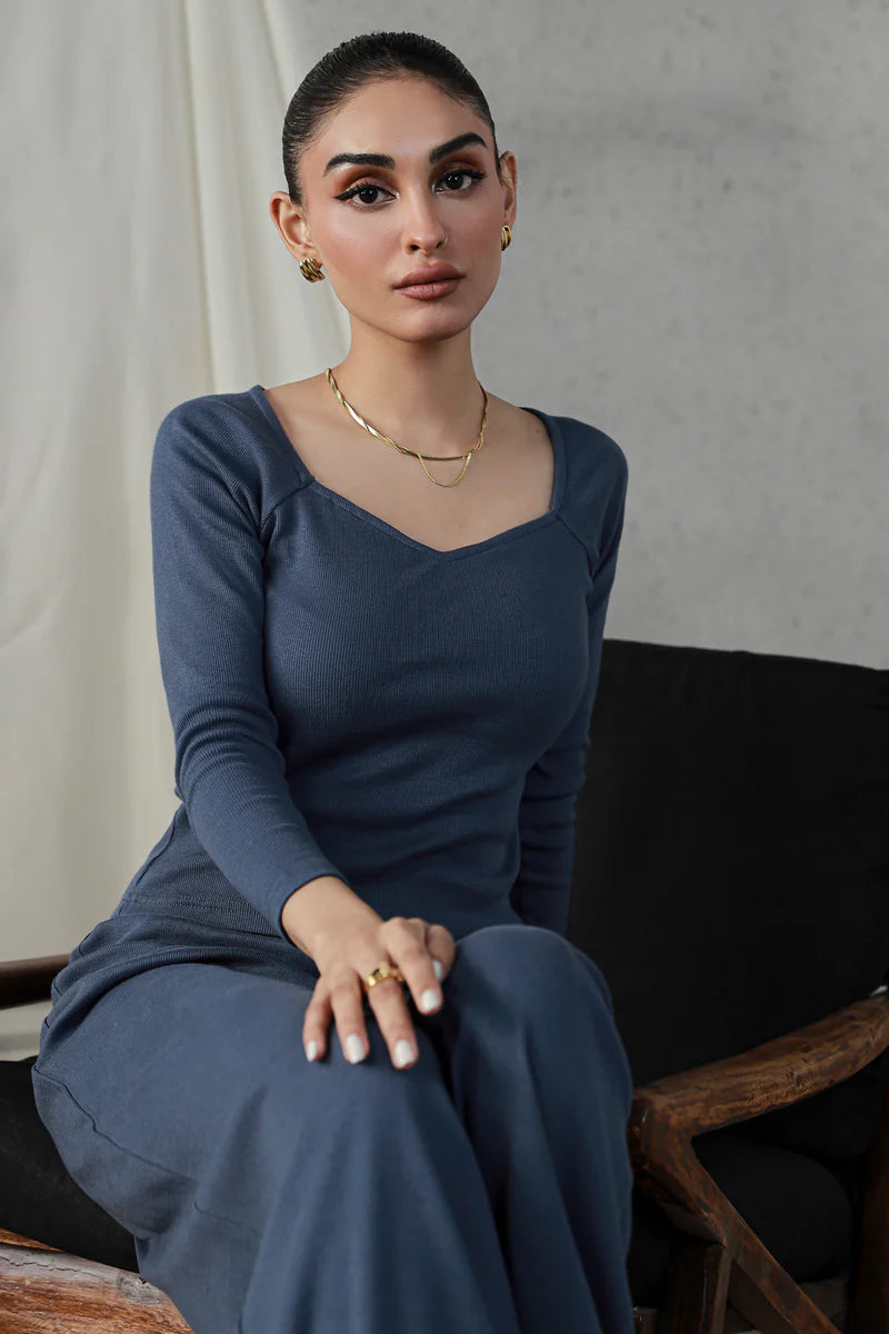 Woman in an indigo sweetheart neck top, accessorized with gold jewelry, seated in a minimalistic setting.